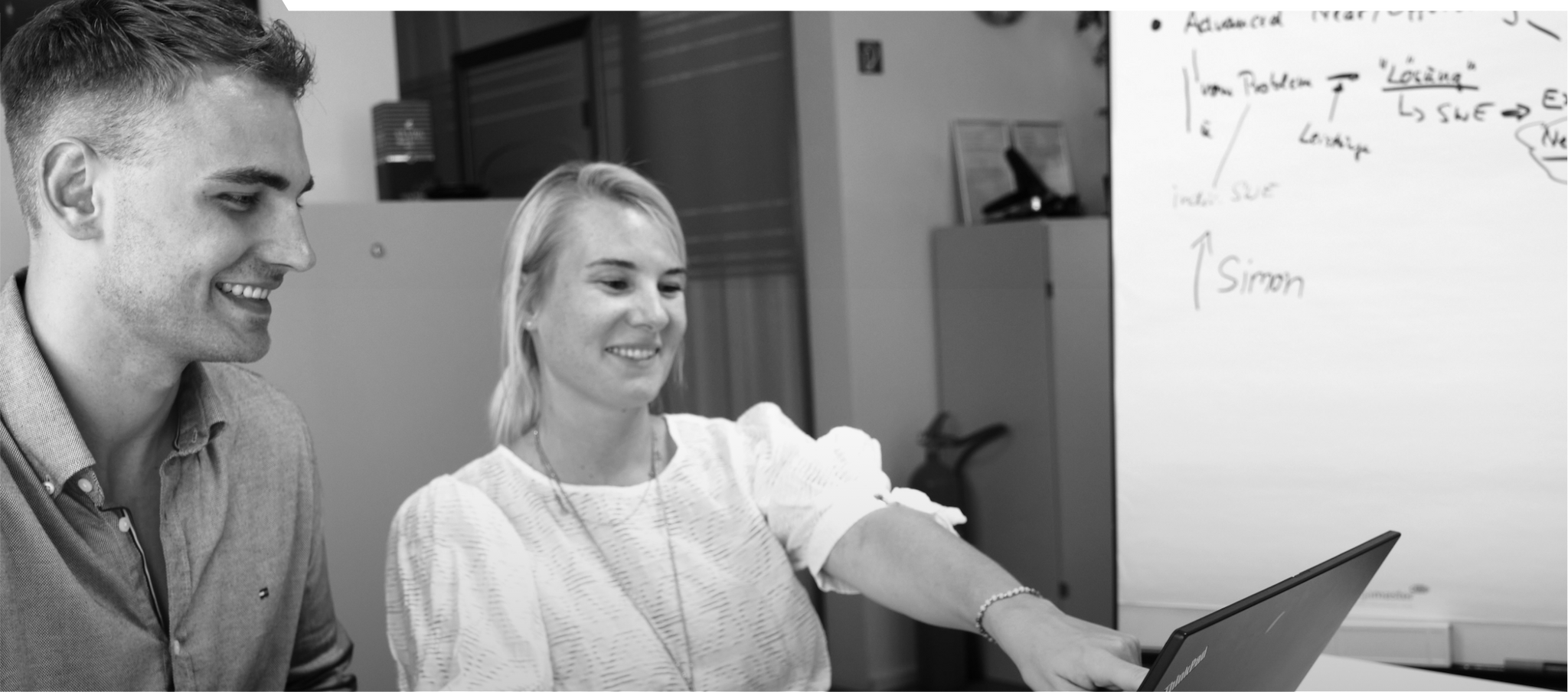 Schwarz-weiß-Bild. Ricarda zeigt auf ihren Laptop und Simon schaut lächeln mit rein in den Laptop.