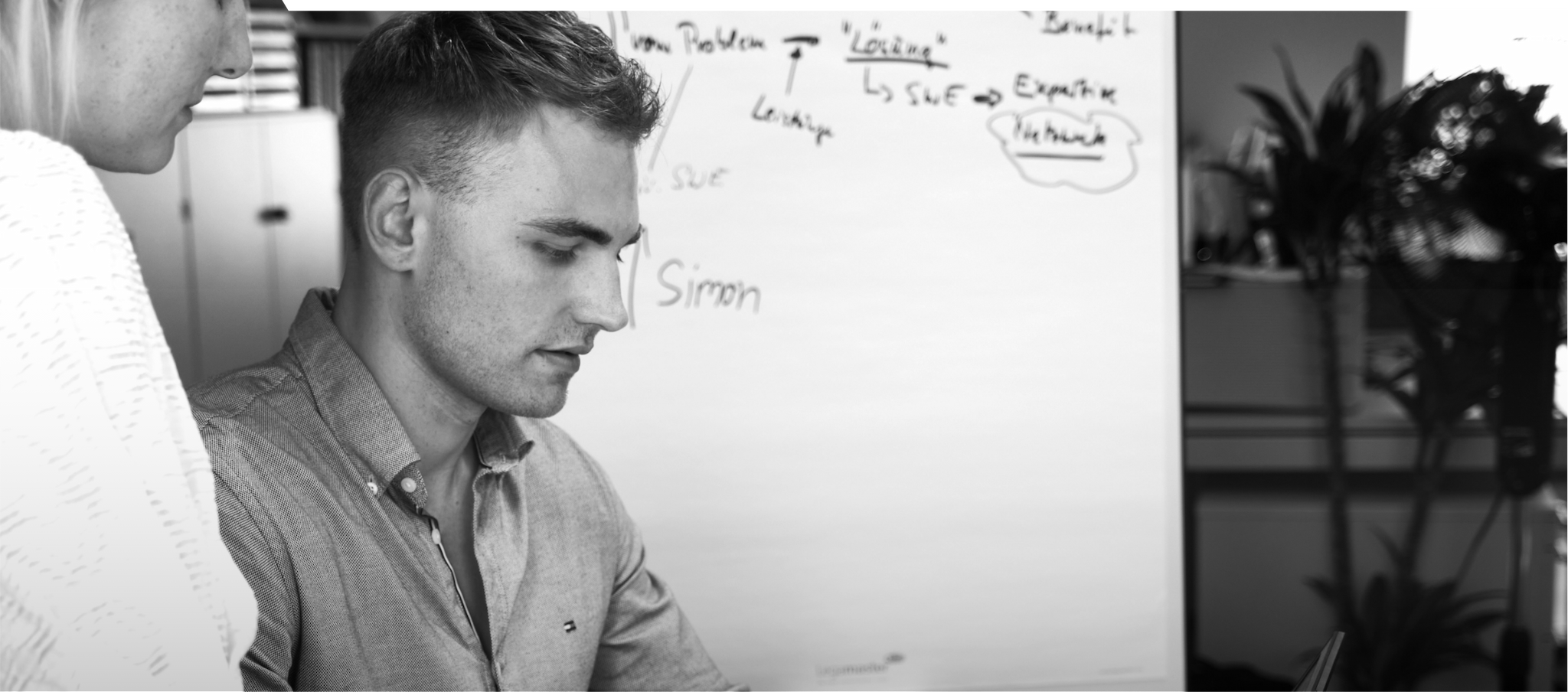 Schwarz-weiß-Bild. Simon schaut konzentriert auf irgendetwas. Im Hintegrund sieht man ein Whiteboard.