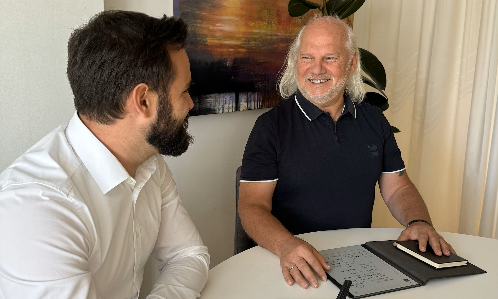Bernd und Fabian schauen sich lächelnd an und sind gerade in einer Besprechung für ServiceFactum. Sie sitzen beide im Besprechungsraum.